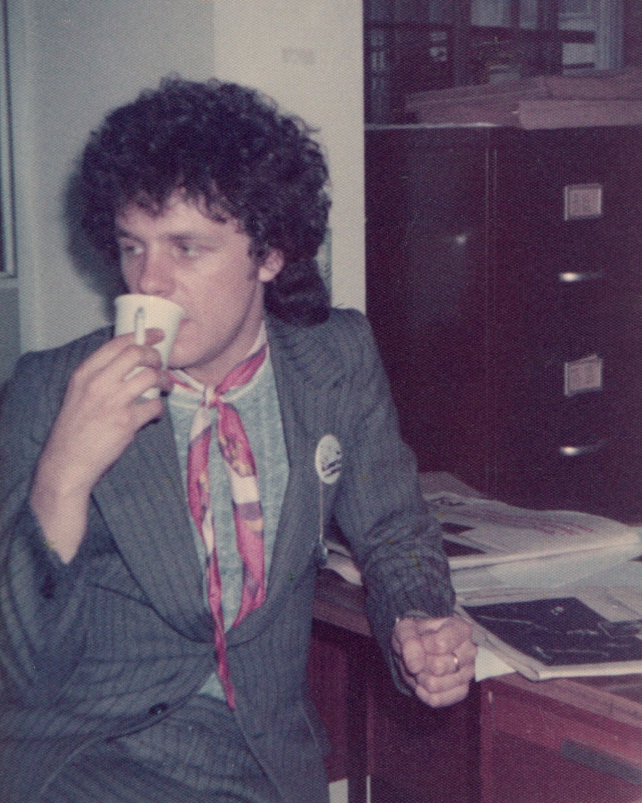 Steve McGarry in a grey pinstripe suit with a pink neckerchief holding a coffee cup and cigarette while wearing his Slaughter and the Dogs badge